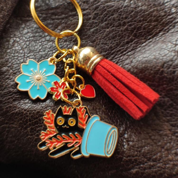 Porte-clé chat noir dans pot bleu renversé avec plante rouge, pompon rouge sur sac marron