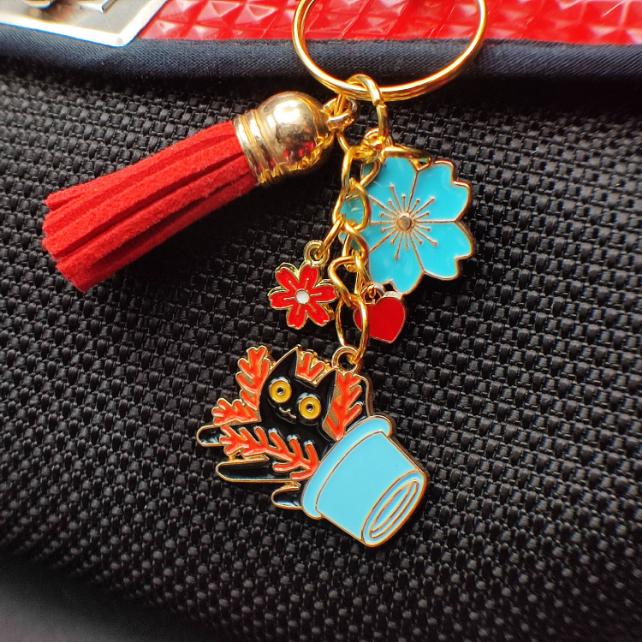 Porte-clé chat noir dans pot bleu renversé avec plante rouge, pompon rouge sur sac noir et rouge