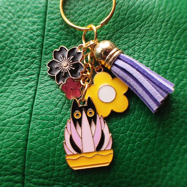 Porte-clé chat noir dans un pot jaune avec plante rose et mauve, pompon mauve sur un sac vert

