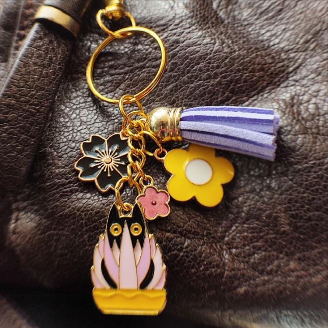 Porte-clé chat noir dans un pot jaune avec plante rose et mauve, pompon mauve sur un sac marron